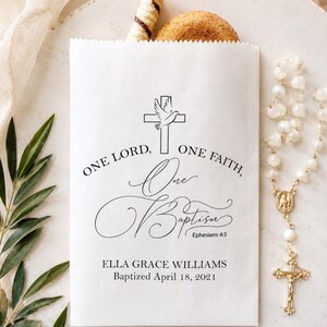 May include: A white paper favour bag featuring black printed text that reads One Lord, One Faith, One Baptism, Ephesians 4:5, Ella Grace Williams, and Baptised April 18, 2021, with a graphic of a cross and a dove above the text.