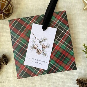 May include: A red, green, and black plaid gift bag with a white gift tag. The gift tag has a watercolor illustration of pine branches with pine cones and the text "season's greetings, the Wilsons".