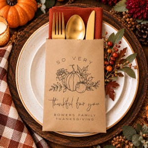 May include: A brown paper cutlery pouch containing a gold fork, spoon, and knife. The pouch features a black line drawing of a pumpkin, leaves, and berries with the text: SO VERY thankful for you BOWERS FAMILY THANKSGIVING, placed on a plate.
