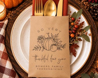 May include: A brown paper cutlery pouch containing a gold fork, spoon, and knife. The pouch features a black line drawing of a pumpkin, leaves, and berries with the text: SO VERY thankful for you BOWERS FAMILY THANKSGIVING, placed on a plate.