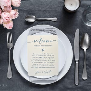 K&ouml;nnte beinhalten: Eine Hochzeitstafel mit einem wei&szlig;en Teller, Besteck und einer Willkommenskarte. Auf der Karte steht "welcome FAMILY AND FRIENDS" mit einem Herzsymbol. Rosa Rosen und eine Kerze sind ebenfalls zu sehen. Die Tischdecke ist dunkelgrau.