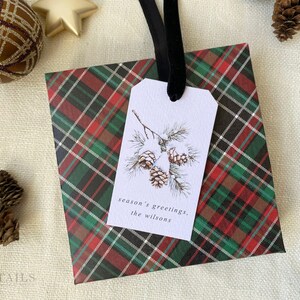 May include: A red, green, and black plaid gift bag with a white gift tag. The tag has a watercolor illustration of pine branches with pine cones and the text "season's greetings, the Wilsons".