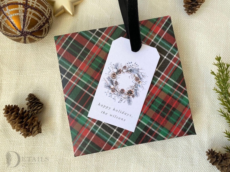May include: A red, green, and black plaid gift bag with a white gift tag. The gift tag has a watercolor illustration of a pine cone wreath and the text "happy holidays, the Wilsons".