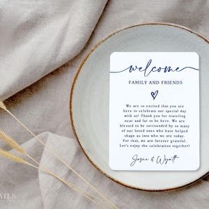 K&ouml;nnte beinhalten: Eine wei&szlig;e rechteckige Karte mit dem Wort "welcome" in marineblauer Schrift, zusammen mit dem Text "FAMILY AND FRIENDS" und einem Herzsymbol. Die Karte liegt auf einem wei&szlig;en Teller mit braunem Rand, auf einer beigen Leinenoberfl&auml;che.