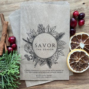 Puede incluir: Una bolsa de papel marrón con una ilustración en blanco y negro de una corona con el texto "Savor the Season". La bolsa también incluye instrucciones para hervir el contenido en la estufa con agua para llenar tu hogar con aromas navideños. La bolsa está etiquetada con "Con amor de la familia Lux."