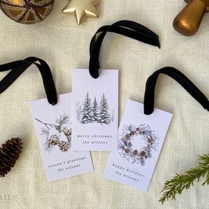 May include: Three white gift tags with black ribbon. The tags feature watercolor illustrations of pine trees, a wreath, and pine cones. The text on the tags reads "merry christmas, the wilsons", "season's greetings, the wilsons", and "happy holidays, the wilsons".