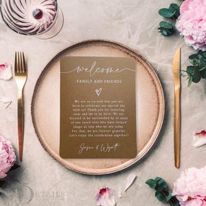 K&ouml;nnte beinhalten: Eine Hochzeitstafel mit goldener Gabel und Messer, einem braunen Teller und einer Karte mit der Aufschrift "Willkommen Familie und Freunde". Rosa Blumen und ein Glas Wein sind ebenfalls vorhanden.