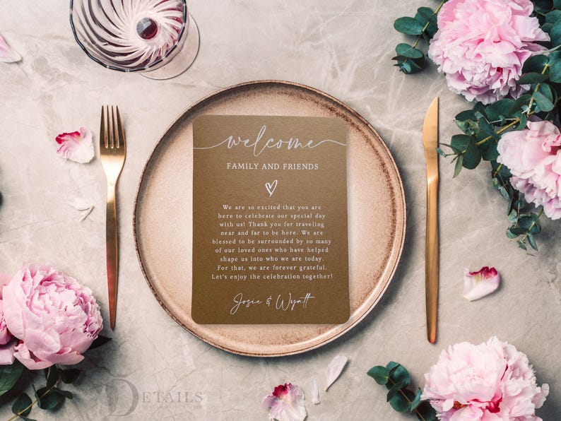 K&ouml;nnte beinhalten: Eine Hochzeitstafel mit goldener Gabel und Messer, einem braunen Teller und einer Karte mit der Aufschrift "Willkommen Familie und Freunde". Rosa Blumen und ein Glas Wein sind ebenfalls vorhanden.