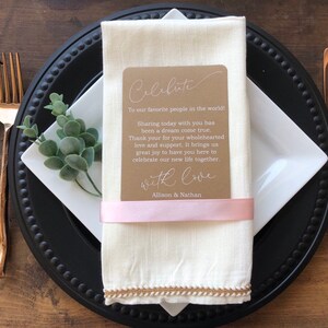 May include: A white linen napkin with a brown card that says "Celebrate" and "To our favorite people in the world! Sharing today with you has been a dream come true. Thank you for your wholehearted love and support. It brings us great joy to have you here to celebrate our new life together. With love, Allison & Nathan". The napkin is tied with a pink ribbon and is on a black charger plate with a white plate on top.