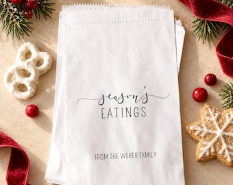 May include: A stack of white paper treat bags featuring the black printed text "season's EATINGS" in a mix of script and sans-serif fonts, followed by the text "FROM THE WEBER FAMILY" in a simple sans-serif typeface at the bottom.