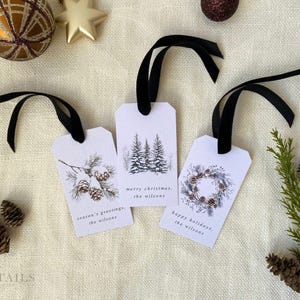 Three watercolor-style Christmas gift tags with black ribbon.  Tags feature winter-themed designs: pine cones, snow-covered trees, and a wreath.  Text on tags includes 'Merry Christmas', 'Season's Greetings', and 'Happy Holidays', along with 'the Wilsons'.  Tags are off-white with dark-green and brown accents.