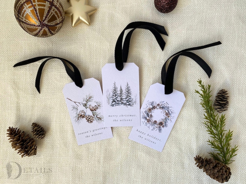 Three watercolor-style Christmas gift tags with black ribbon.  Tags feature winter-themed designs: pine cones, snow-covered trees, and a wreath.  Text on tags includes 'Merry Christmas', 'Season's Greetings', and 'Happy Holidays', along with 'the Wilsons'.  Tags are off-white with dark-green and brown accents.