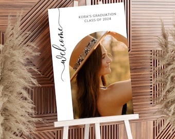 May include: White welcome sign with black calligraphy. The sign has a photo of a young woman wearing a brown and black hat. The text on the sign reads "Kora's Graduation Class of 2024".