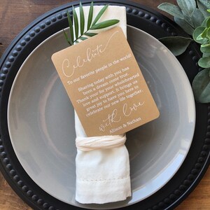 May include: A white napkin with a brown card attached to it. The card has white text that reads "Celebrate" and "To our favorite people in the world! Sharing today with you has been a dream come true. Thank you for your wholehearted love and support. It brings us great joy to have you here to celebrate our new life together. With love, Allison & Nathan".