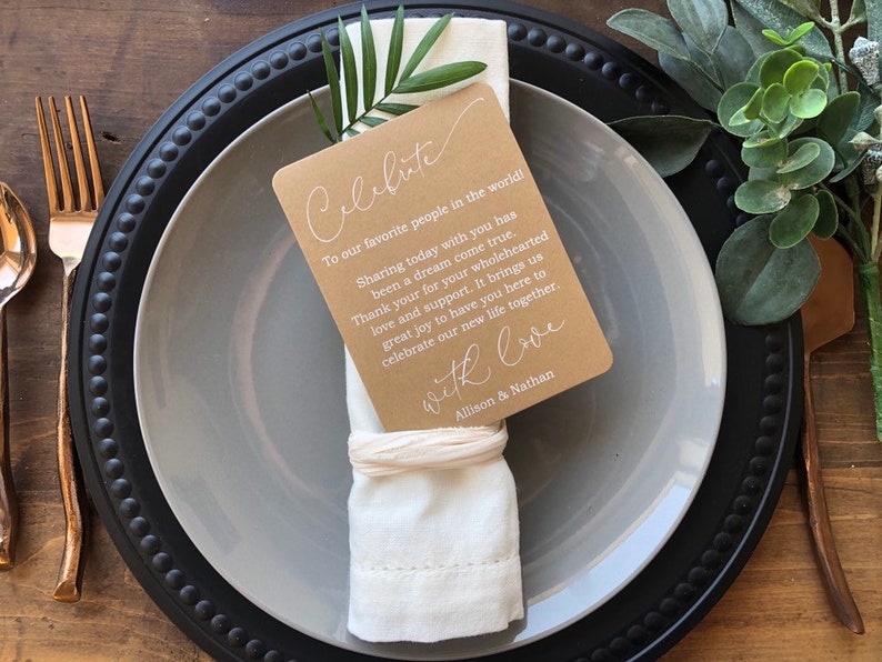 May include: A white napkin with a brown card attached to it. The card has white text that reads "Celebrate" and "To our favorite people in the world! Sharing today with you has been a dream come true. Thank you for your wholehearted love and support. It brings us great joy to have you here to celebrate our new life together. With love, Allison & Nathan".