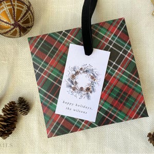 May include: A red and green plaid gift bag with a white gift tag. The gift tag has a watercolor illustration of a pine cone wreath and the text "happy holidays, the Wilsons".