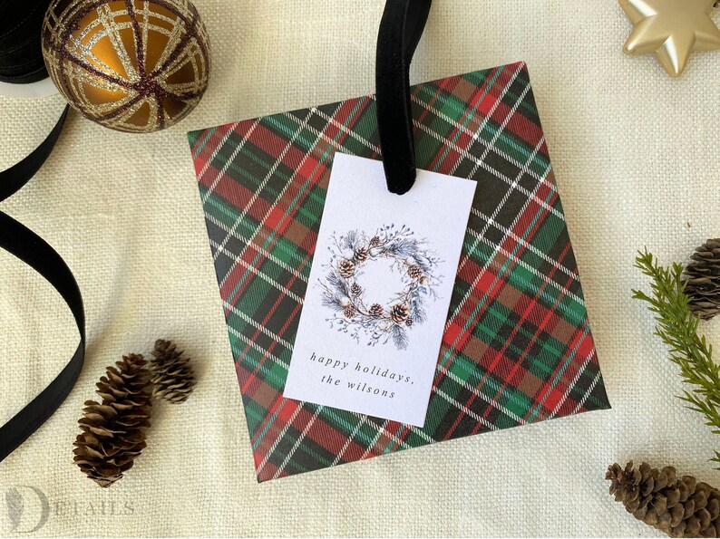 May include: A red and green plaid gift bag with a white gift tag. The gift tag has a watercolor illustration of a pine cone wreath and the text "happy holidays, the Wilsons".