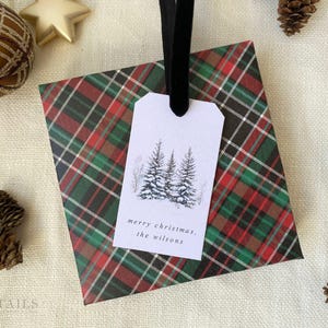 May include: A white gift tag with a black and white illustration of three pine trees with snow on the branches. The tag is tied with a black ribbon and is on top of red and green plaid wrapping paper. The text on the tag reads "merry christmas, the wilsons".