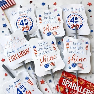 May include: A collection of white sparkler tags with patriotic designs and text. The tags feature phrases like "Ignite the light and let it shine" and "Baby, you're a FIREWORK." An American flag and star-shaped confetti are also visible.