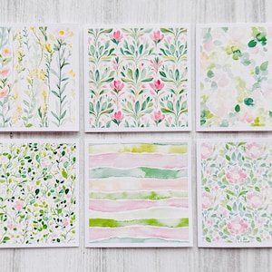 May include: Six square watercolor art prints featuring floral and abstract designs. The prints showcase a variety of patterns, including delicate flowers, leafy branches, and watercolor stripes, in soft pastel colors like pink, green, and yellow.