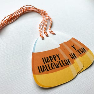 May include: Three orange and yellow candy corn shaped gift tags with twine attached. The tags have the text "Happy Halloween!" printed on them.