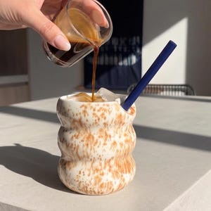 May include: A wavy, cream-coloured ceramic cup with brown speckles, filled with ice cubes. A dark blue straw rests inside the cup. A clear glass vessel is pouring dark brown espresso into the cup, creating a layered coffee beverage.