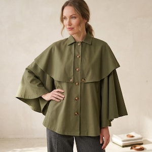 May include: An olive green cape jacket featuring a structured collar, a button-down front with brown buttons, and a wide, layered cape-style overlay that extends over the shoulders and arms, creating a distinct, flowing silhouette.