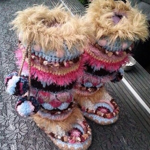 May include: A pair of mid-calf crochet boots featuring a multi-textured design with layers of pink, blue, brown, and cream yarn. The boots are topped with tan faux fur trim and include decorative yarn pom-poms hanging from the side.