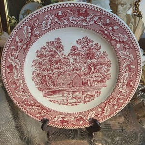 May include: A round ceramic plate featuring a red and white transferware pattern. The center depicts a rural landscape with a house, trees, and figures near a pond. The rim is decorated with a detailed border of oak leaves and acorns.