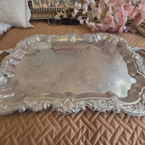 May include: A rectangular silver-toned serving tray featuring ornate, raised floral and scrollwork detailing along the scalloped edges and two integrated handles. The central surface displays a subtle, etched pattern across the metal.