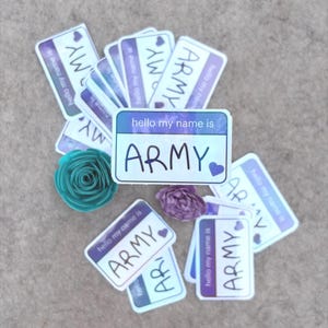 Puede incluir: Una colección de pegatinas rectangulares con un diseño en morado y blanco con el texto hello my name is ARMY y un pequeño corazón morado. Dos pequeñas flores de papel en turquesa y morado están colocadas entre las etiquetas adhesivas.