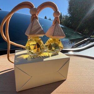 May include: Two glass car air freshener bottles with faceted geometric shapes, filled with a yellow liquid. Each bottle features a light-colored wooden pyramid-shaped cap and a matching beige hanging cord. They sit atop a beige box labeled FRESHENER.
