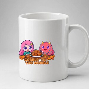 May include: A white ceramic mug featuring a colourful cartoon illustration of a pink-haired character and a pink monster sitting at a table with a roasted turkey, pumpkins, and autumn leaves, with the text GOBBLE 'TIL YOU WOBBLE printed below.