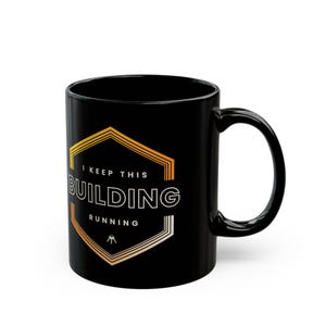 May include: A black ceramic mug featuring a graphic design of a hexagon with orange and yellow gradient lines. The text reads I KEEP THIS BUILDING RUNNING in white capital letters, with a small icon of three keys at the bottom centre.