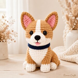 May include: A crocheted Corgi dog toy featuring tan and white yarn, large black plastic eyes, a black nose, pink inner ears, a small pink tongue, and a dark blue collar. The stuffed animal is sitting upright with white paws and a white chest.