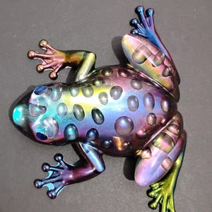 May include: A frog figurine featuring an iridescent, multi-colored finish that shifts between shades of blue, purple, yellow, and green. The surface is covered in raised, circular bumps across the back and head, with detailed toes.