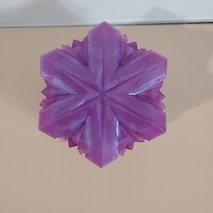 May include: A six-pointed star-shaped object in a vibrant purple hue with a subtle pearlescent finish. The surface features geometric, faceted edges that radiate from the center, creating a sharp, symmetrical, and crystalline appearance.