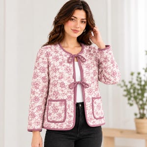 May include: A quilted jacket featuring a light pink background with a repeating dark pink floral pattern. The jacket has long sleeves, a round neckline, two front patch pockets, and two tie closures at the center front with dark pink trim.