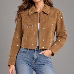 May include: A tan suede jacket featuring a collared neckline, a front zip closure, and a horizontal zipped chest pocket. The jacket is decorated with numerous small, circular silver-toned metal studs scattered across the front and sleeves.