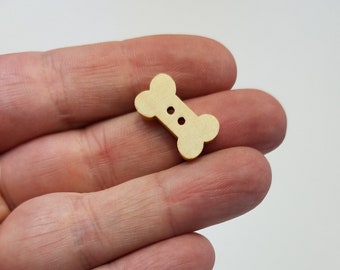 May include: A close-up of a wooden bone-shaped button with two holes. The button is light brown and is being held in a hand. There are several other bone-shaped buttons in the background.