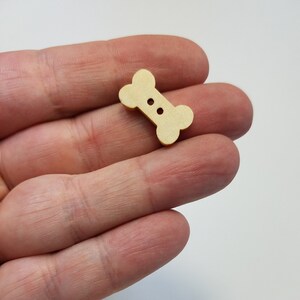May include: A close-up of a wooden bone-shaped button with two holes. The button is light brown and is being held in a hand. There are several other bone-shaped buttons in the background.