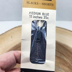May include: A vintage Talon zipper for trousers, slacks, or shorts. The packaging is tan with black text and a blue zipper. The text on the package reads "MEDIUM BLUE 11 inches 35c" and "fly placket zipper."