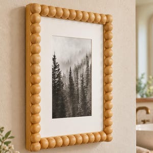 May include: A rectangular wall frame featuring a light-toned wooden border composed of a series of connected spheres. The frame contains a white mat surrounding a black and white photograph depicting a dense forest landscape shrouded in thick fog.