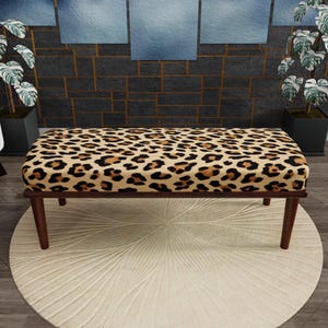 May include: A rectangular bench featuring a tan, brown, and black leopard print upholstered seat. The bench is supported by four dark brown wooden legs that taper towards the bottom, providing a stable base for the cushioned top surface.