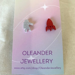 May include: Space-themed stud earrings. One earring is a silver glitter astronaut, the other a red glitter rocket. The earrings are displayed on a card with the text "OLEANDER JEWELLERY" and a website address.