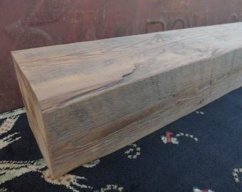 May include: A rustic wooden beam with a natural, weathered finish. The beam has a rectangular shape and is made of rough-hewn wood with visible knots and grain patterns. The wood is a light brown color with streaks of gray and white.