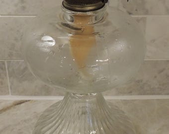 May include: A clear glass oil lamp with a ribbed base and a metal burner. The lamp is sitting on a tiled surface.