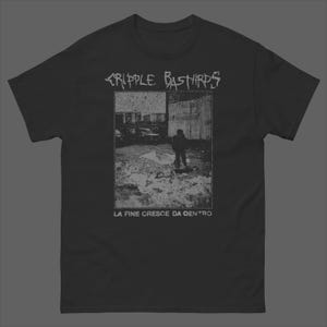 以下が含まれることがあります： 白いグラフィックプリントが施された黒の半袖Tシャツ。上部に「CRIPPLE BASTARDS」、下部に「LA FINE CRESCE DA DENTRO」というテキストが配置され、都市環境にいる人物の粒子状のモノクロ画像が中央に描かれています。