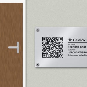 Könnte beinhalten: Ein rechteckiges silbernes Metallschild mit vier Eckschrauben, das einen schwarzen QR-Code sowie den Text Gäste-WLAN, Netzwerk Seeblick-Gast, Passwort Sonnenschein2026 und Code scannen und verbinden neben einer braunen Tür zeigt.