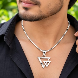 May include: A silver-toned necklace featuring a box chain and a pendant composed of two overlapping open triangles. The pendant has a polished metallic finish and hangs from a simple bail attached to the chain, resting against a black collared shirt.
